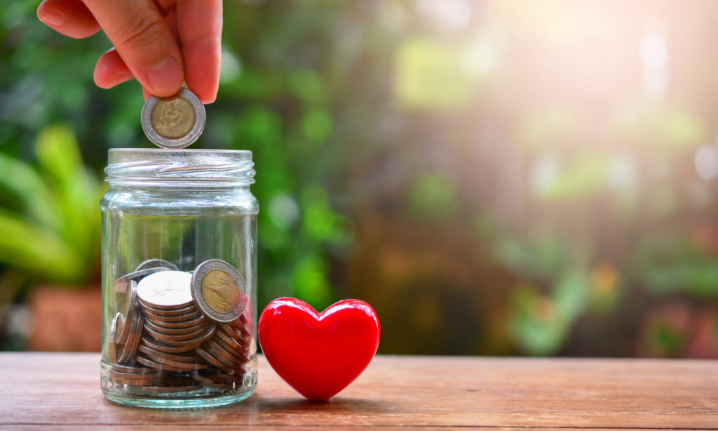 Pote de vidro com moedas dentro e uma mão com uma moeda, com um coração do lado e fundo de jardim